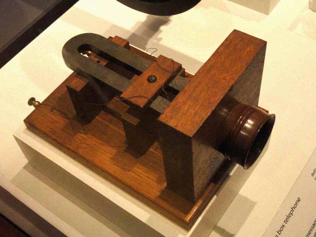 A wooden box telephone with a metal receiver and coil mounted on top, displayed on a white platform in a museum setting.