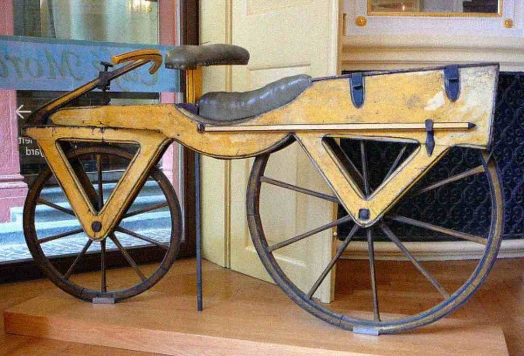 A vintage, yellow, wooden bicycle with triangular frame and large spoked wheels is displayed indoors near a window. The bike has no pedals and features a leather seat.