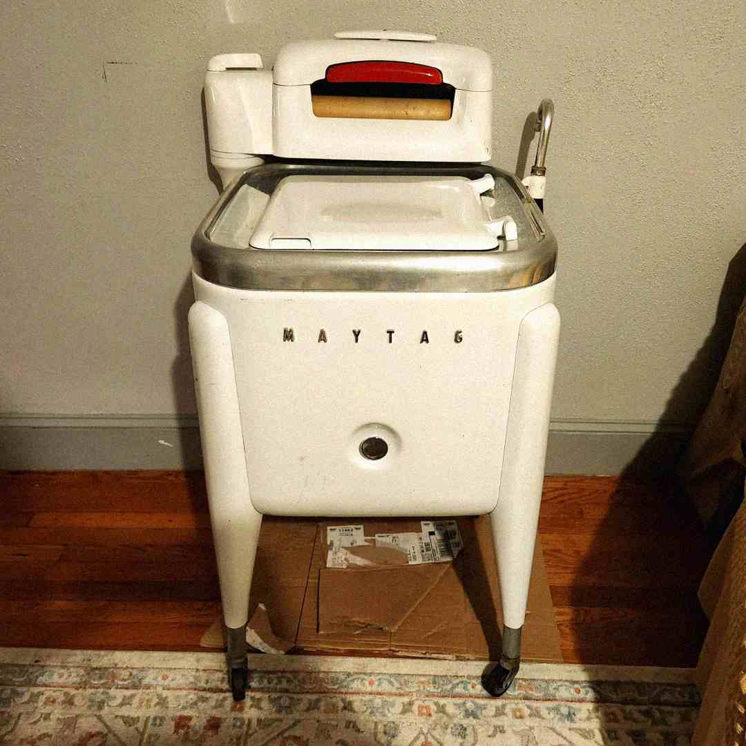 A vintage white Maytag washing machine with a wringer on top, standing on four legs with wheels, placed on a cardboard piece over a hardwood floor near a wall and a patterned rug.