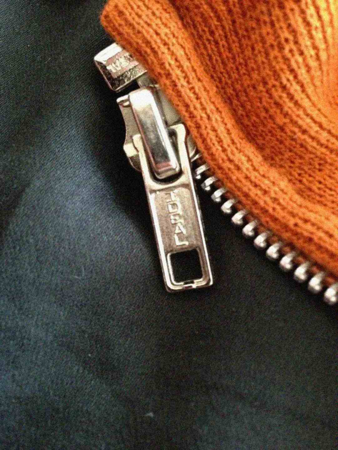 A close-up of a metallic zipper on a garment, with the zipper pull labeled "IDEAL." The zipper is attached to an orange fabric, and the surrounding material is black.