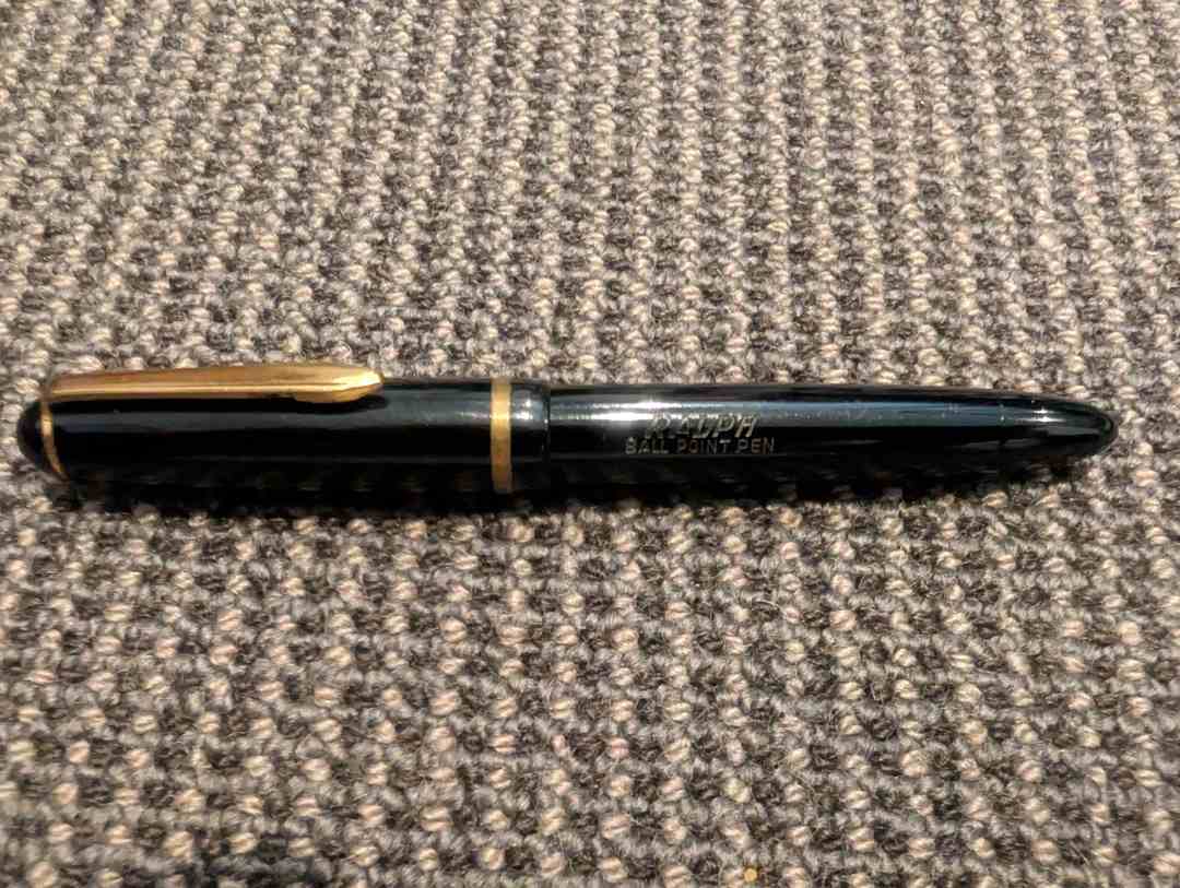 A black ballpoint pen with gold accents lies horizontally on a textured gray carpet. The pen cap is on, and the gold clip and band are visible.