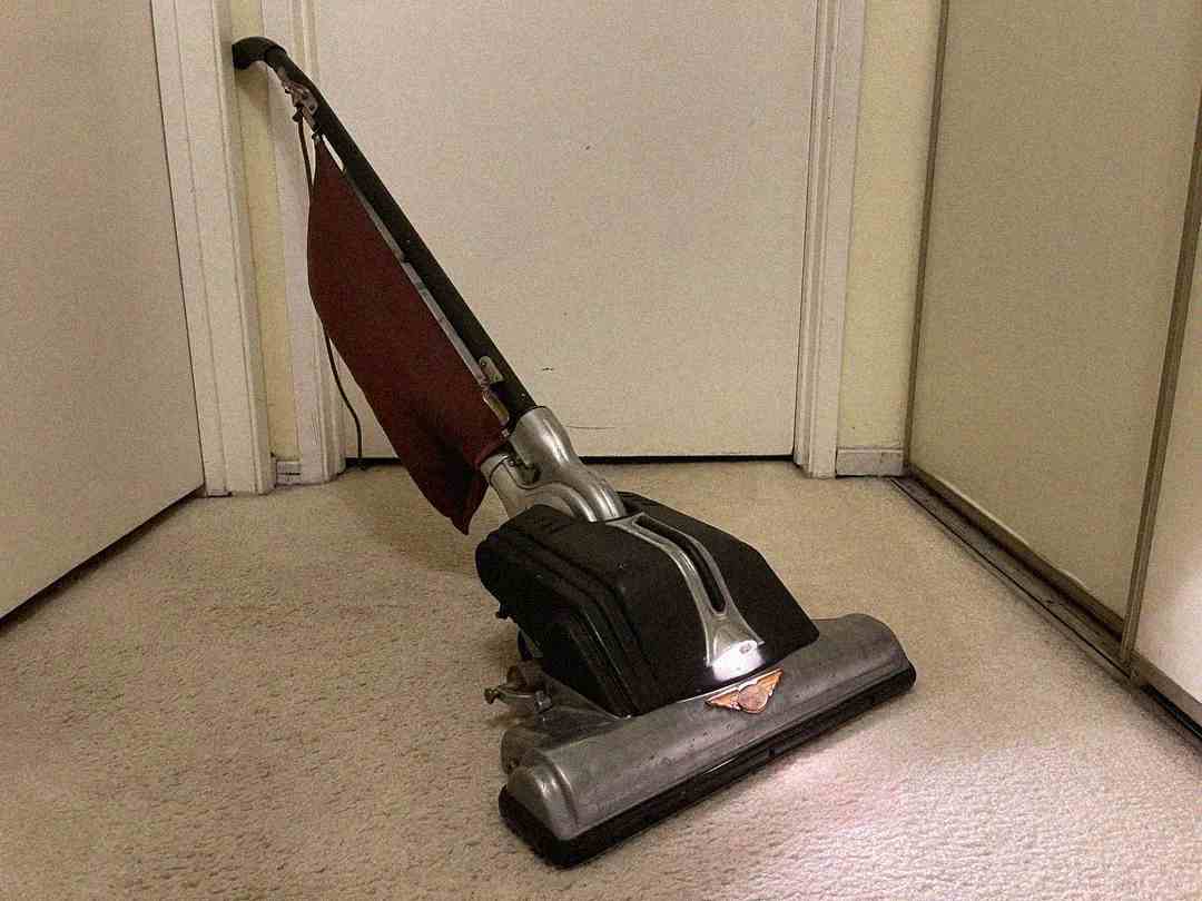 A vintage upright vacuum cleaner with a red fabric dust bag stands on a beige carpet in a corner, surrounded by white doors and walls.