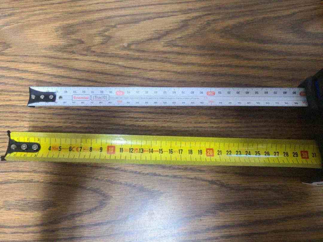 Two tape measures are extended side by side on a wood-patterned surface. The top tape is silver with black and red markings, while the bottom tape is yellow with black and red markings. Both show centimeter measurements.