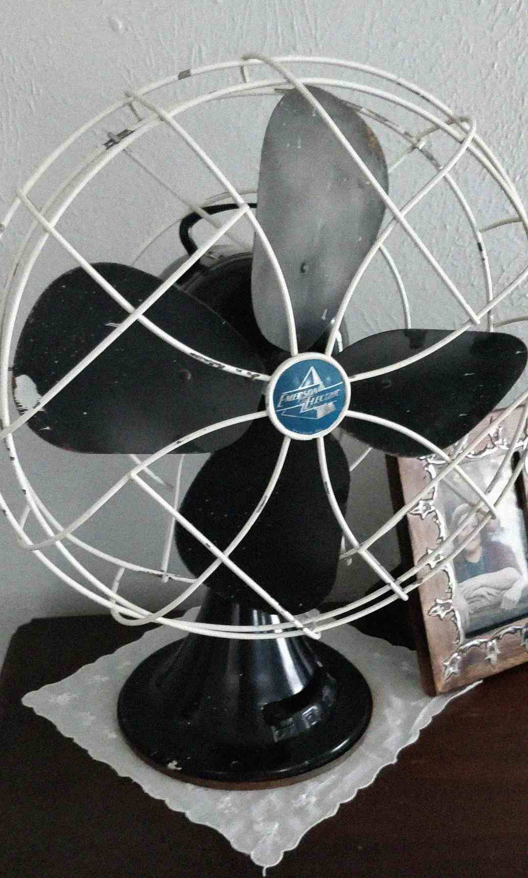 A black vintage electric table fan with a metal cage sits on a dark surface next to a framed photo, all resting on a white doily. The background is a plain light-colored wall.