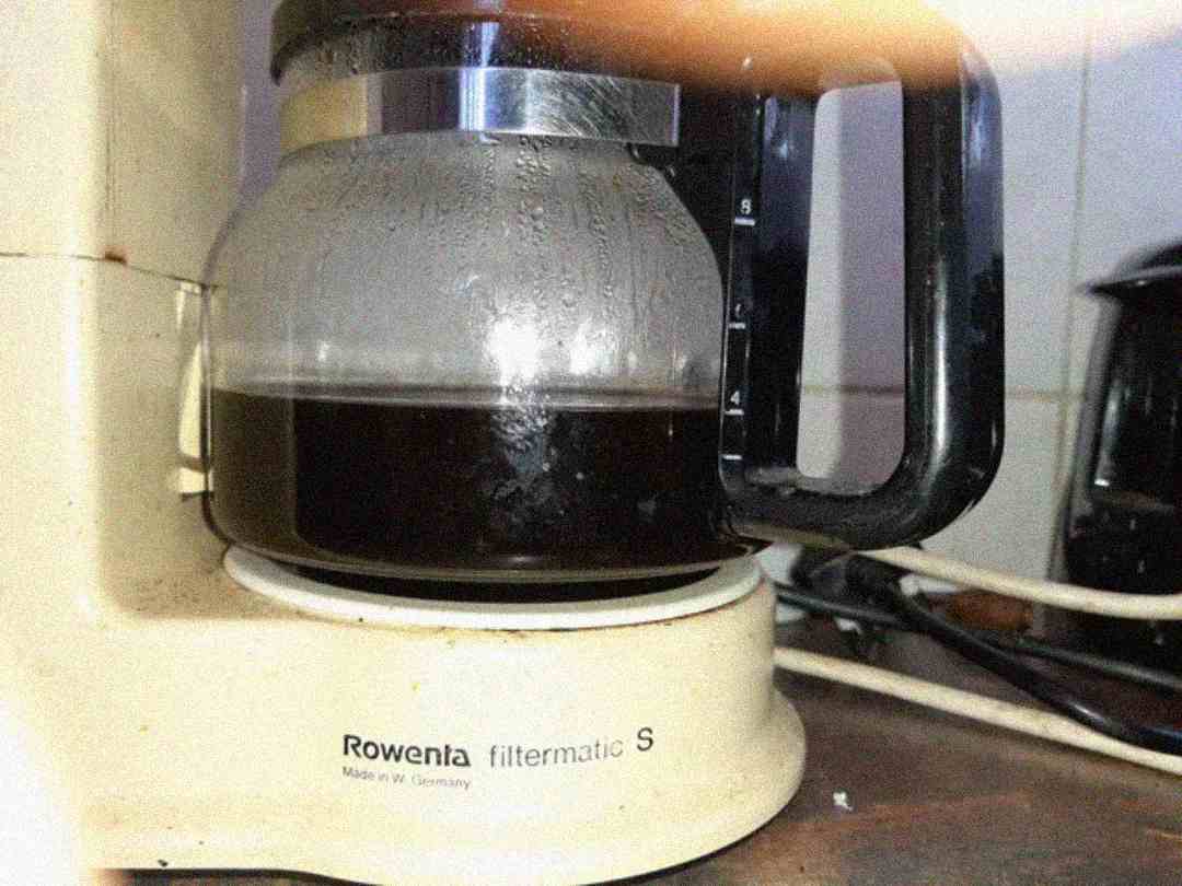 A close-up of a Rowenta filtermatic S coffee maker with a glass pot containing partially brewed black coffee. The machine is beige and sits on a kitchen counter with visible cords in the background.