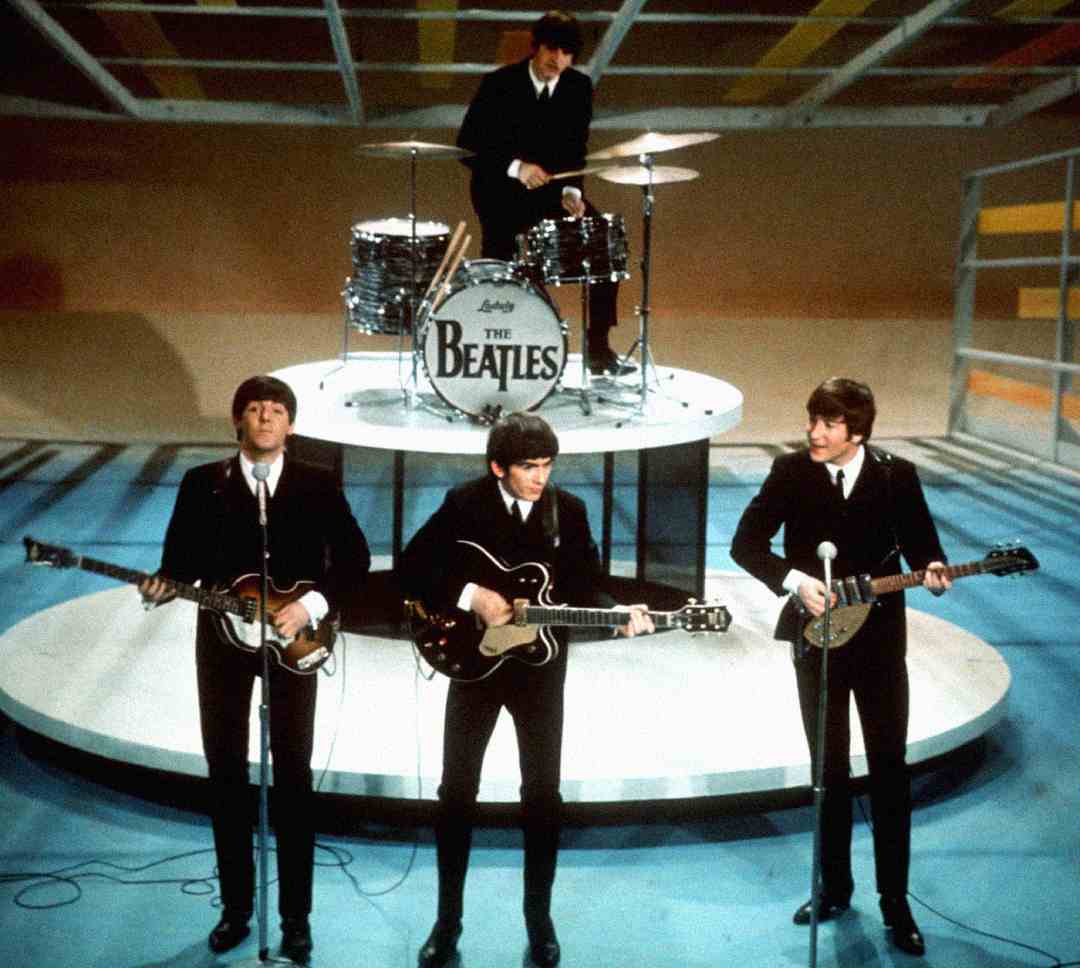 Four musicians in matching black suits perform onstage. Three play guitars and sing, while one plays drums elevated behind them. The drum set displays "The Beatles" logo. The background is colorful and modern.