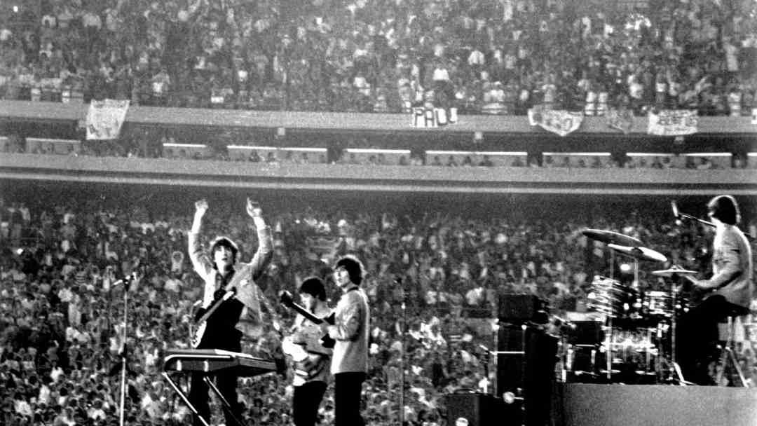 A black-and-white photo of a band performing on stage in a large stadium, with four musicians facing a huge, cheering crowd. Banners and signs are visible in the stands behind them.