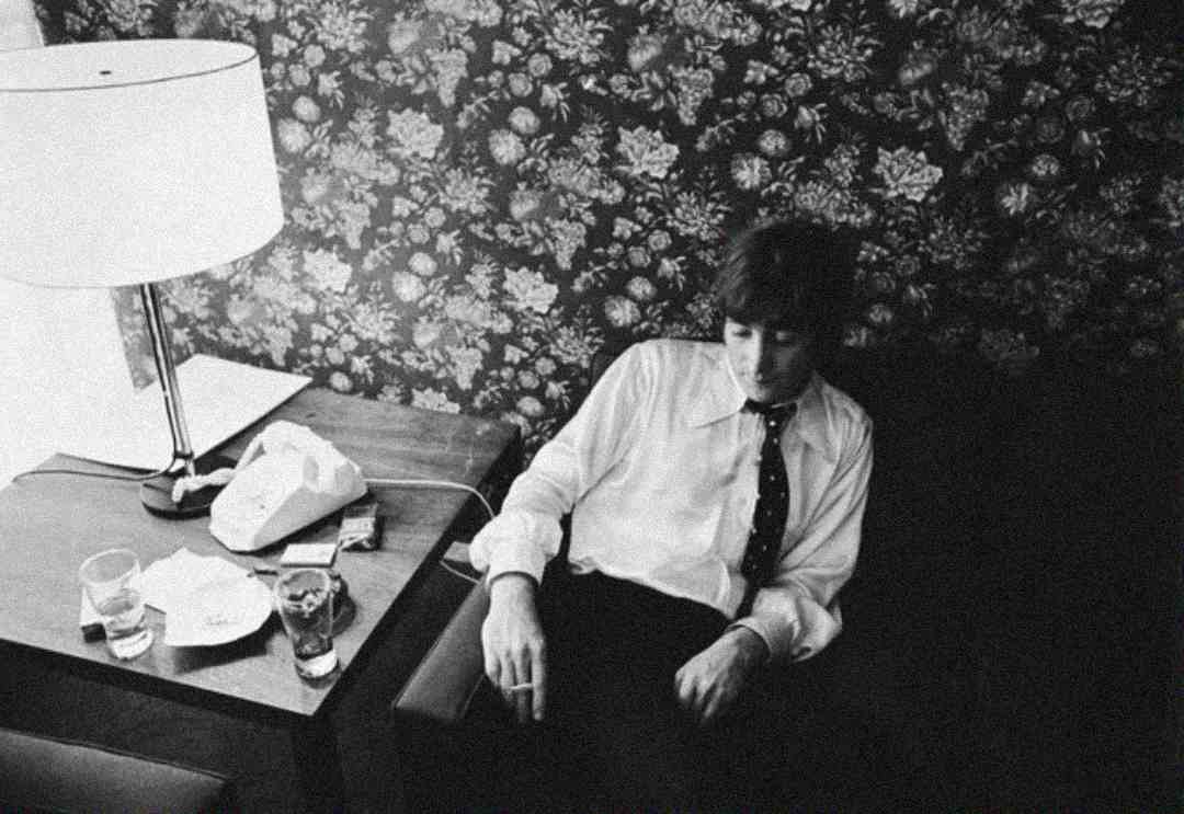 A person in a white shirt and dark tie sits on a couch next to a small table with a lamp, drinks, cigarettes, and a phone. The background features floral wallpaper. The scene is in black and white.