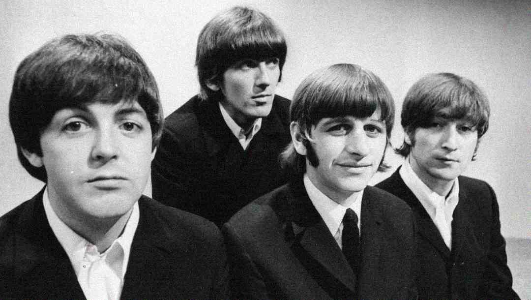 Four young men with dark hair, wearing suits and ties, sit closely together and look toward the camera in a black-and-white image with a plain background.