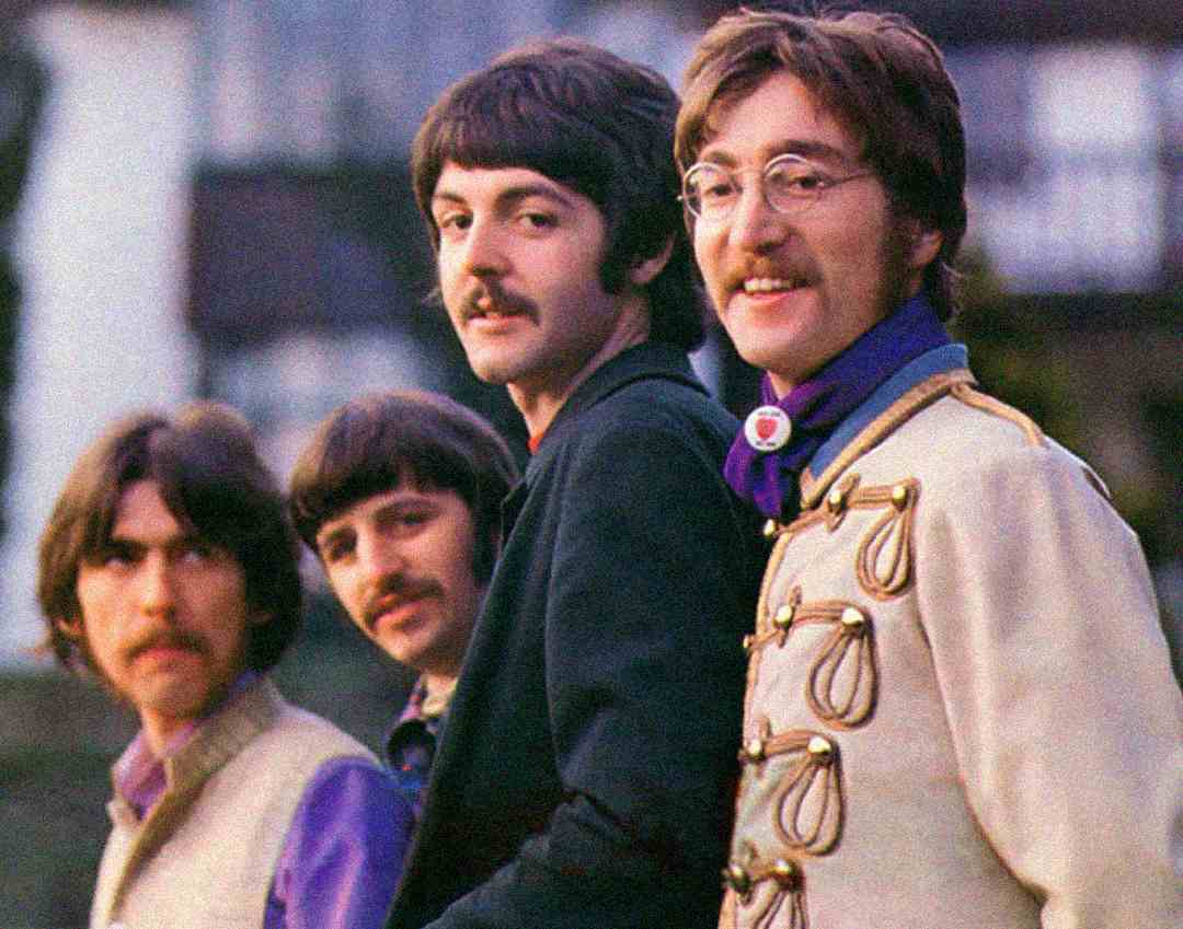 Four men with mustaches, dressed in colorful, vintage-style clothing, stand outdoors. Three look toward the camera while one looks off to the side. The background is blurred, showing greenery and part of a building.
