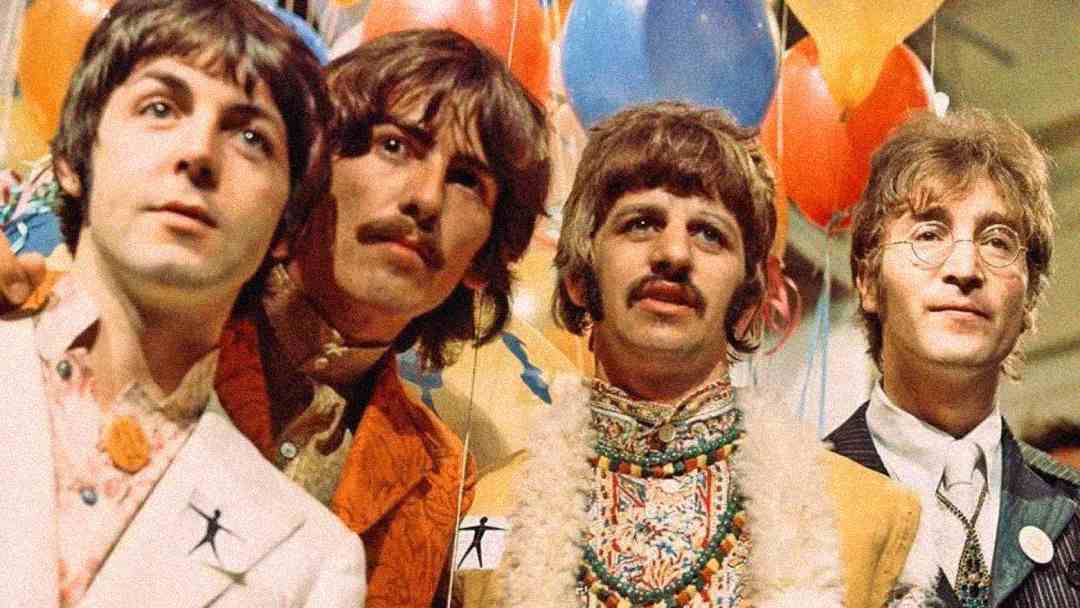 Four men stand closely together in colorful, vintage clothing, surrounded by bright balloons. They have long hair and are looking in different directions, appearing festive and cheerful.