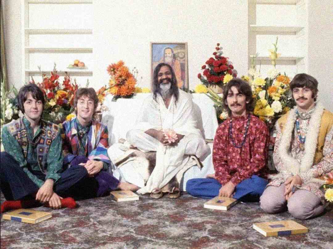 Five men sit cross-legged on the floor in front of flowers and a portrait, with four wearing colorful clothes and one in white robes seated in the center. Each has a book in front of them, and they all smile at the camera.