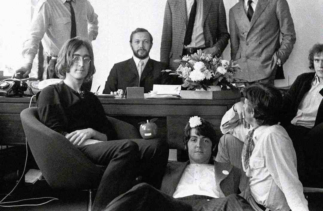 A group of men in suits and casual clothing gather in an office around a desk with flowers; one man sits in a chair, two sit on the floor, and others stand behind the desk. The scene looks informal and relaxed.