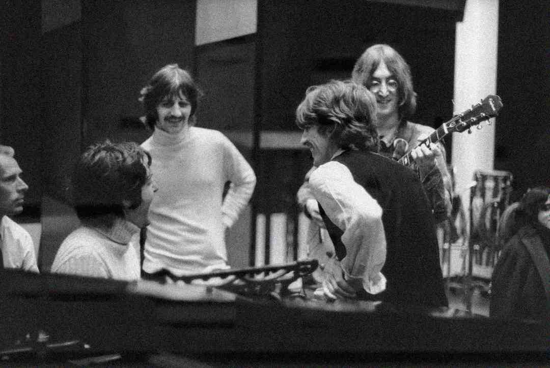 Four men smile and talk together in a recording studio, surrounded by musical equipment. One holds a guitar, while the others are gathered closely, appearing to enjoy a lighthearted moment during a music session.