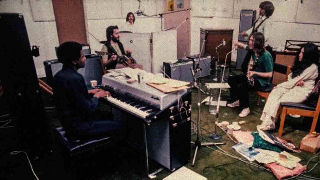 Five musicians sit and play instruments in a cluttered studio, surrounded by papers, cables, amplifiers, and cups. One person sits at an electric piano while others play guitar, drums, and bass, with a woman seated nearby.