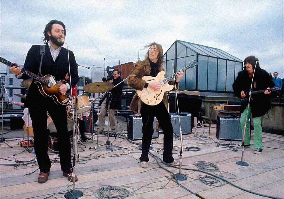 Four musicians perform on a rooftop with instruments including guitars and drums. They are surrounded by microphones and audio equipment, with a glass structure and cloudy sky in the background.