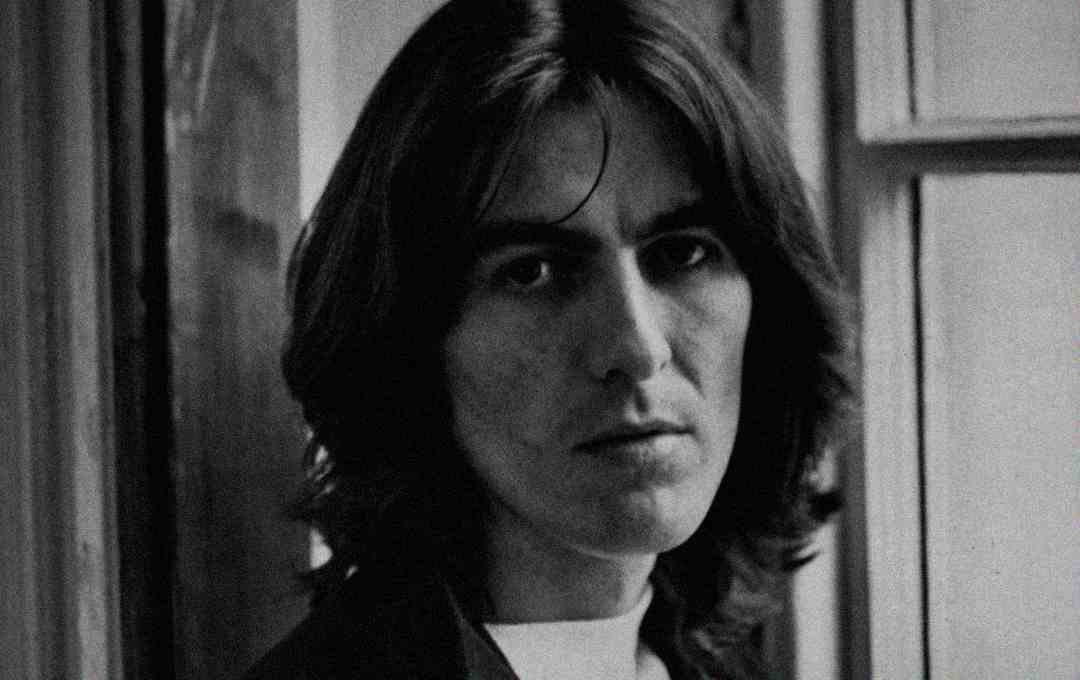 A black-and-white portrait of a young man with shoulder-length dark hair, gazing seriously at the camera, standing beside a window.