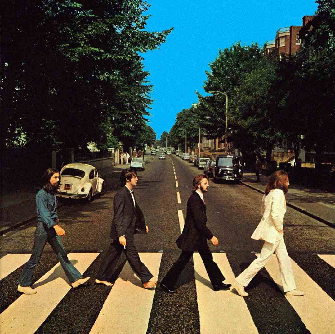 Four men walk barefoot across a zebra crossing on a tree-lined street under a bright blue sky, with a white car parked on the left side of the road.