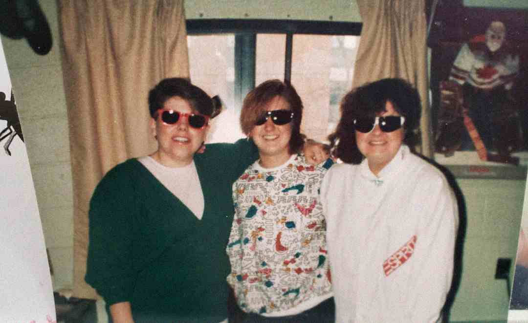 Three people stand indoors in front of a window with curtains, all wearing sunglasses and smiling. Two wear white tops, one with a colorful print, and the third wears a green cardigan. A sports poster hangs on the wall behind them.