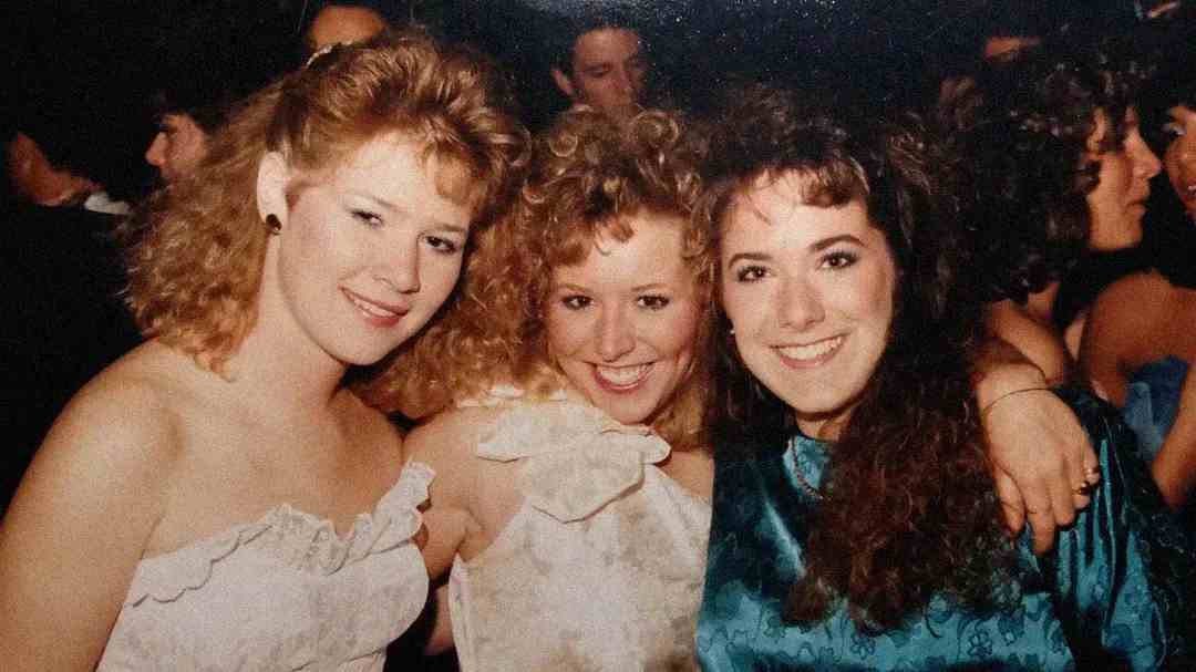 Three young women in 1980s-style dresses smiling and posing together at a crowded party or dance, with curly hair and bright expressions, surrounded by people in the background.