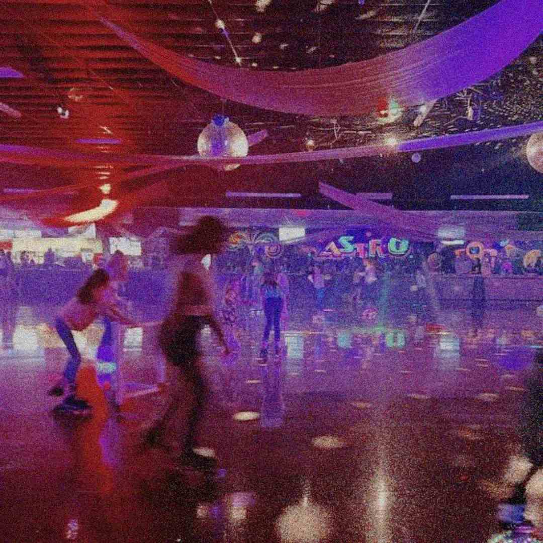 People roller skate under colorful lights and disco balls in a retro-style roller rink. The scene is lively and filled with reflections on the shiny floor. Draped fabric decorates the ceiling, and arcade games line the back wall.