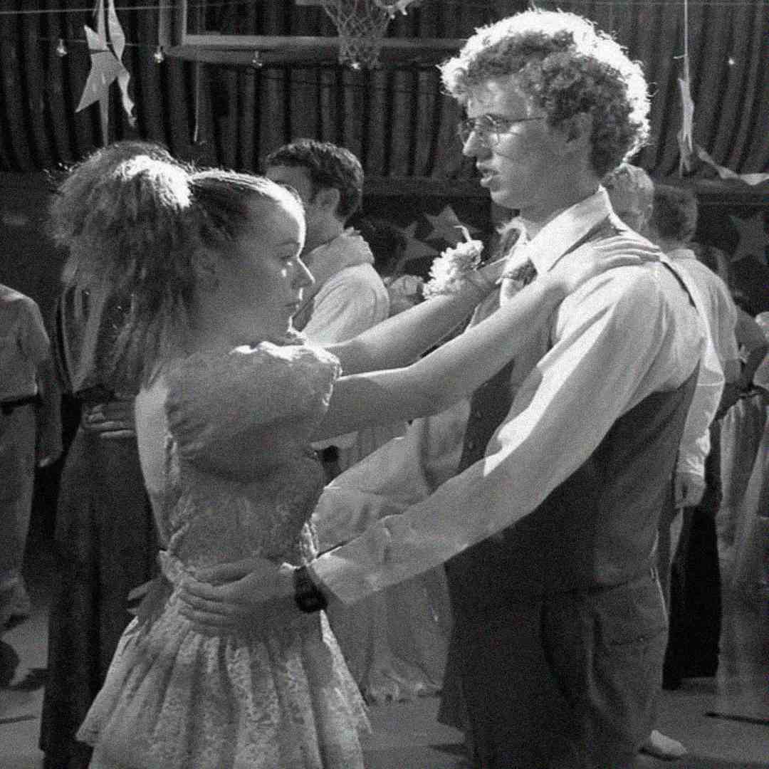 A young couple in formal attire dances closely at a decorated event, with the girl wearing a dress and the boy in a vest and tie. Other people are dancing in the background. The scene is in black and white.