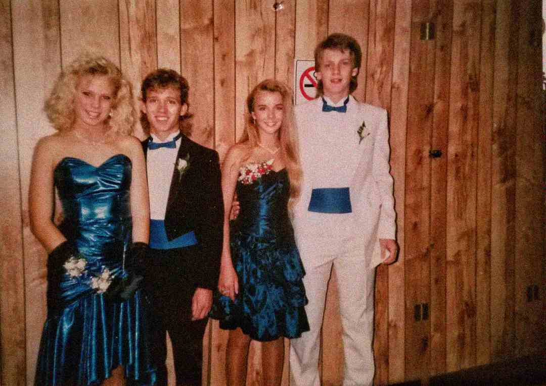 Four teenagers dressed in formal prom attire from the 1980s stand in front of a wood-paneled wall. The two girls wear shiny blue dresses, and the two boys wear tuxedos, one in black and one in white, with matching blue cummerbunds.