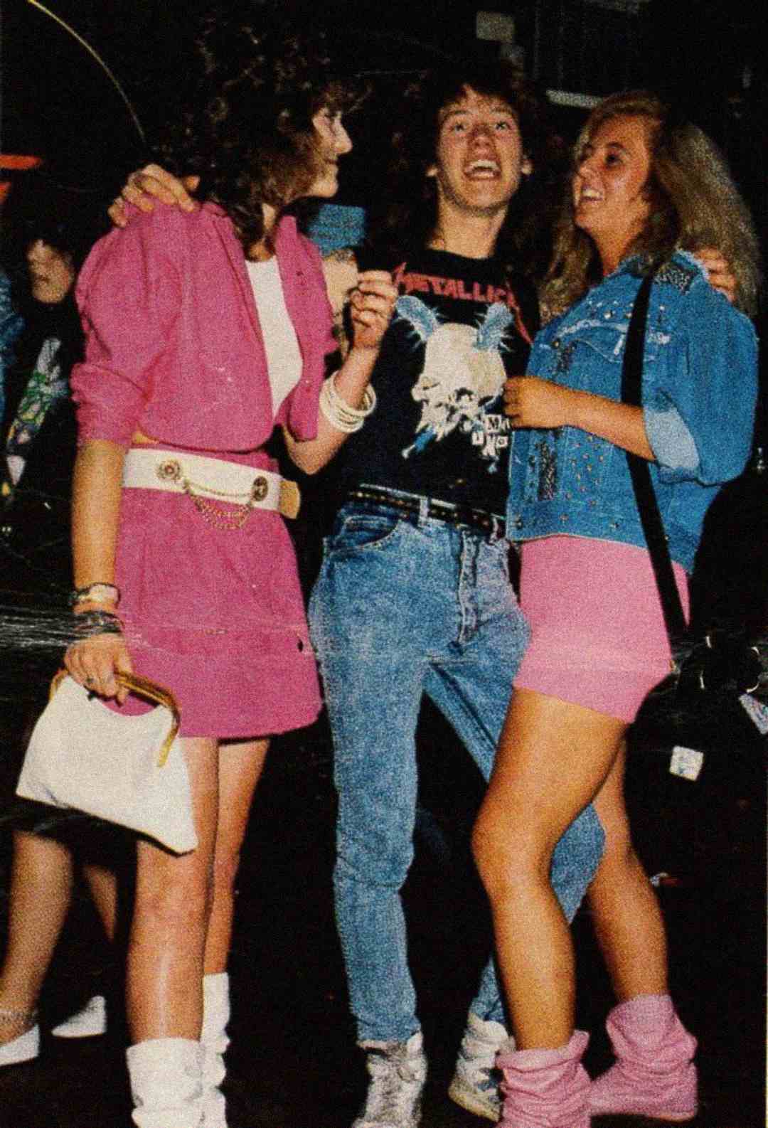 Three young people smile and pose together; two women wear bright 1980s outfits with pink skirts and jackets, and the man in the middle wears a Metallica T-shirt and jeans. They appear to be at a party or club.