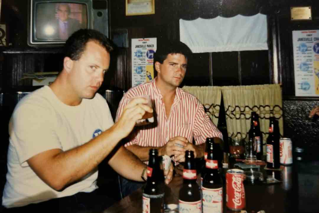 Two men sit at a table with several bottles of Budweiser beer and a can of Coca-Cola. One man in a white shirt holds a drink, while the other in a striped shirt looks ahead. A TV and posters are visible in the background.
