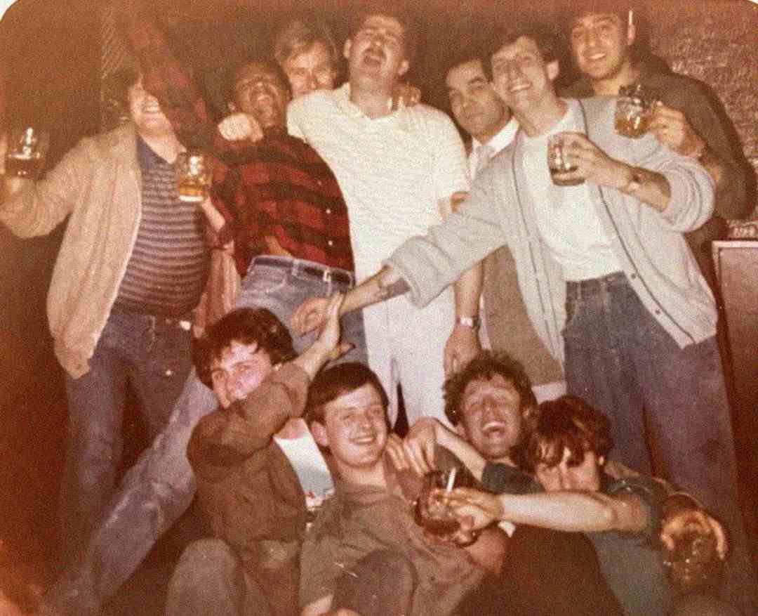 A group of ten men smile, laugh, and pose together while holding drinks, some with arms raised, appearing to celebrate or enjoy a night out in a casual setting.