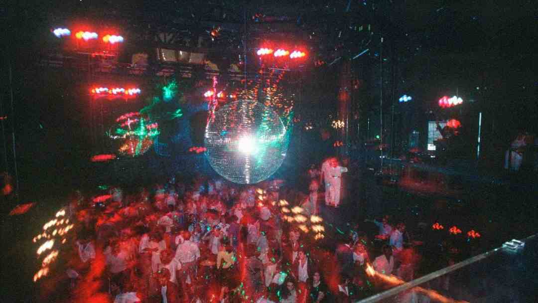 A crowded nightclub dance floor with colorful lights and a large disco ball overhead, creating a lively and energetic atmosphere. The image is slightly blurred from motion.