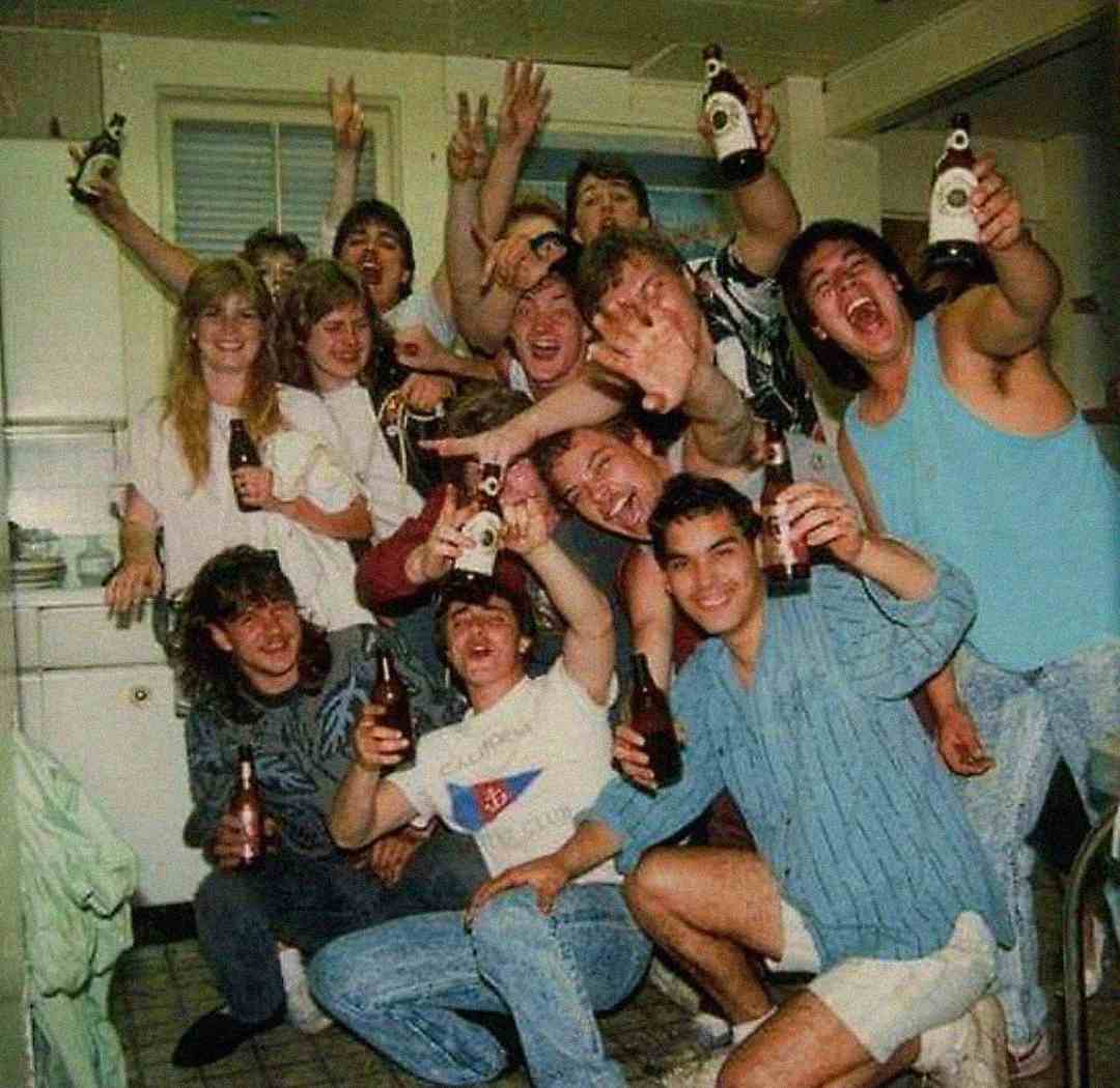 A group of young adults crowded together in a kitchen, smiling, laughing, and holding beer bottles, celebrating and posing energetically for the camera. The atmosphere appears lively and festive.
