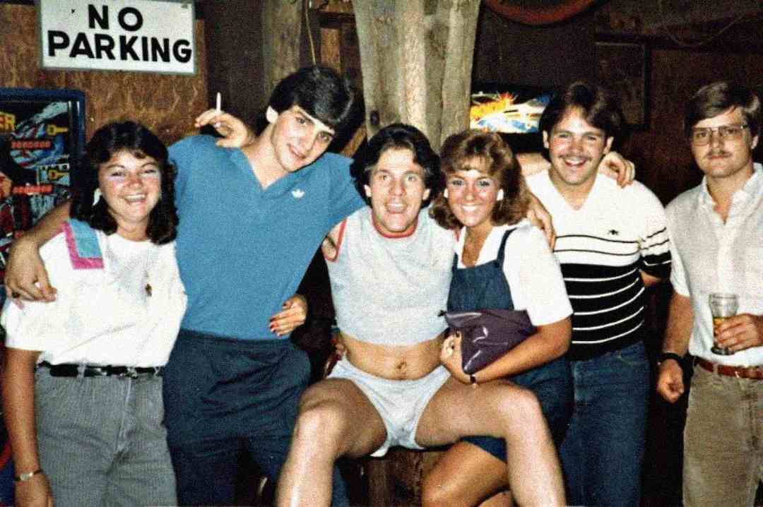 A group of six young adults pose together inside a bar. One man is sitting on a bar, wearing only a T-shirt and underwear. Others stand close, smiling; a "No Parking" sign is visible in the background.