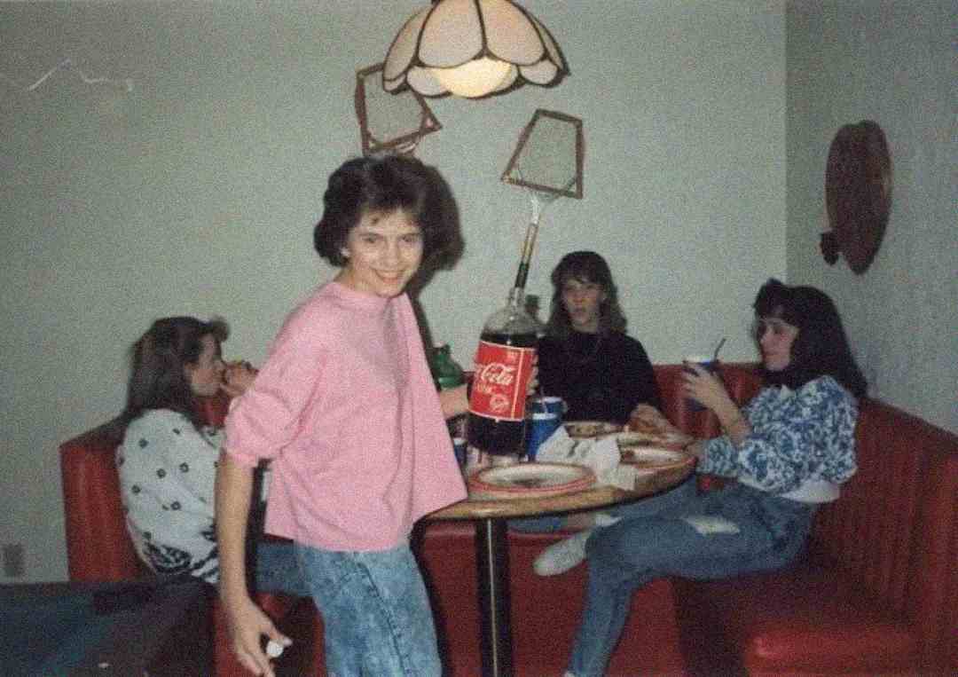 Four young women sit around a dining table with pizza and drinks. One woman in front, smiling, holds a large bottle of Coca-Cola. The room has a red booth seat and a decorative hanging lamp.