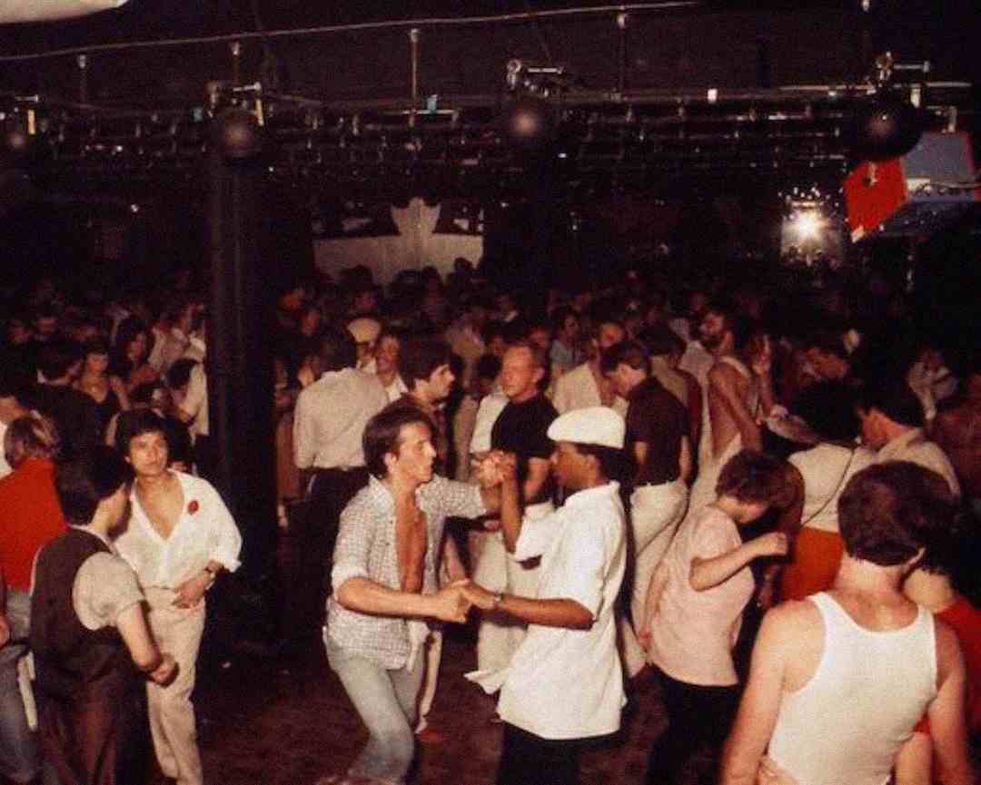 A crowded dance floor with people dancing energetically under dim club lights. Two men are in the foreground holding hands and dancing, surrounded by others enjoying the lively atmosphere.