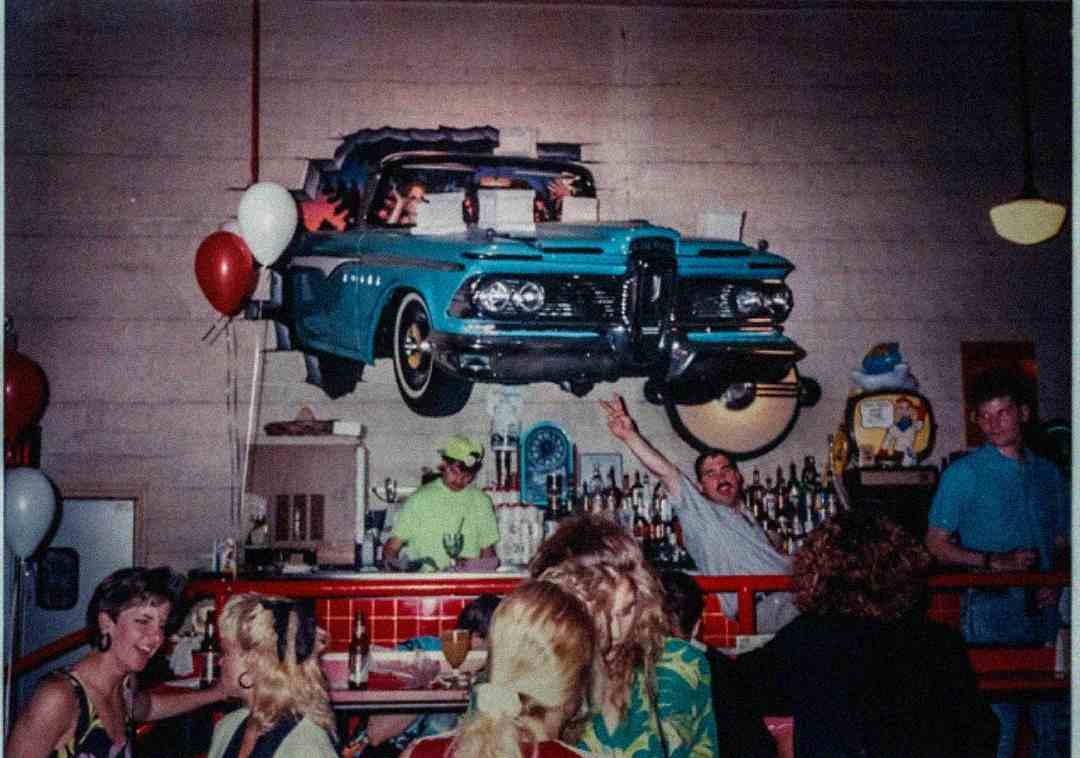A group of people socializing at a bar decorated with a vintage blue car mounted on the wall. Balloons, bottles, and bar decor are visible, creating a lively and retro atmosphere.