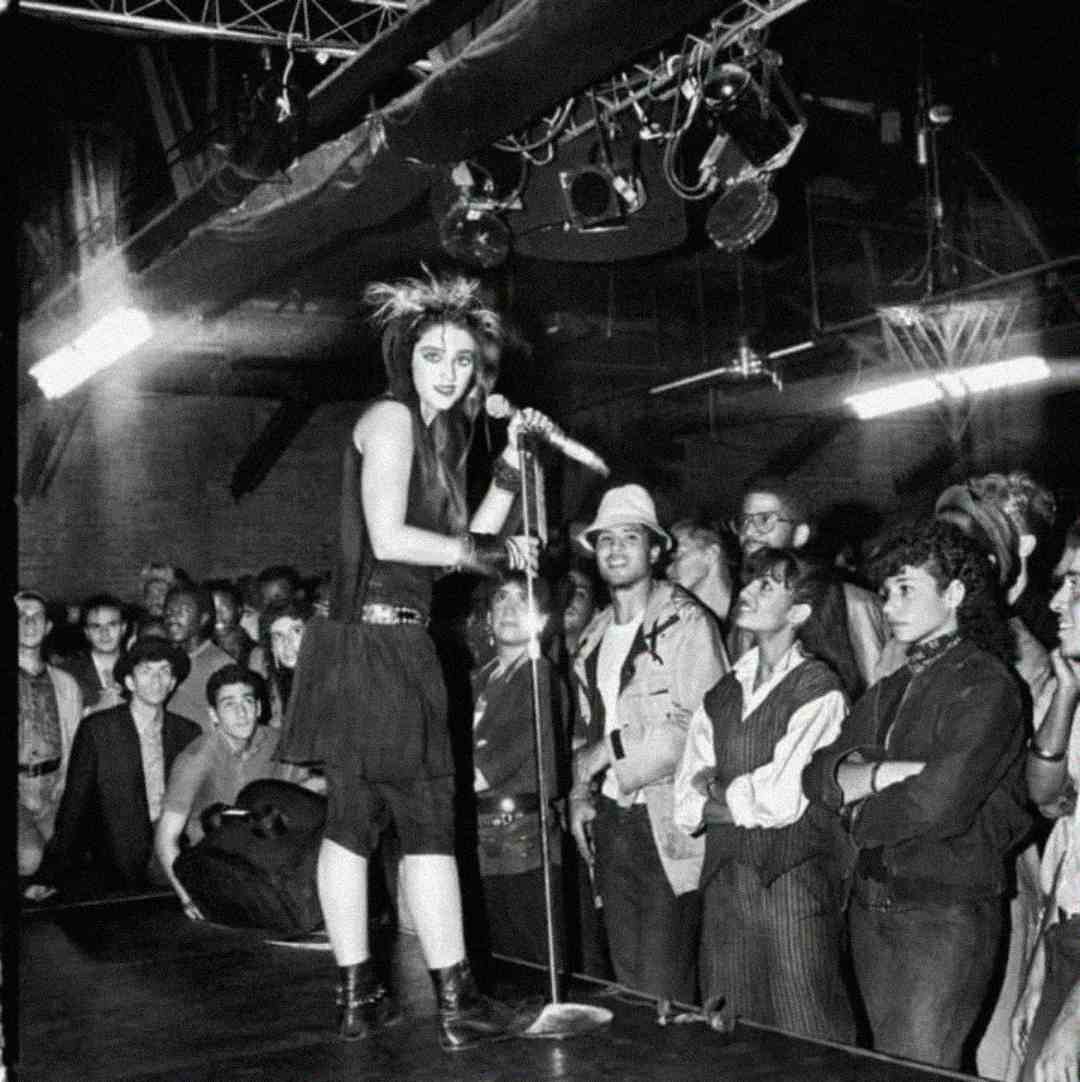 A woman with spiky hair and a dark outfit sings on stage before a crowd in a dimly lit club, as audience members watch her with interest.