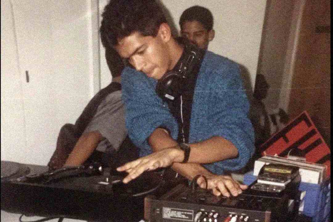 A young man wearing headphones and a blue sweater is DJing, using a turntable and mixer, with another person in the background. Several equipment pieces and a cassette tape are visible on the table.