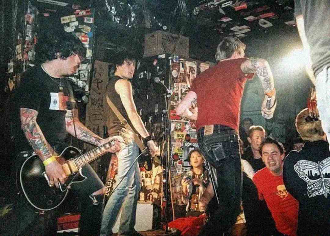 A tattooed punk rock band performs energetically on a small, graffiti-covered stage, while an enthusiastic crowd watches up close. Band members play guitar and sing under colorful stage lights.