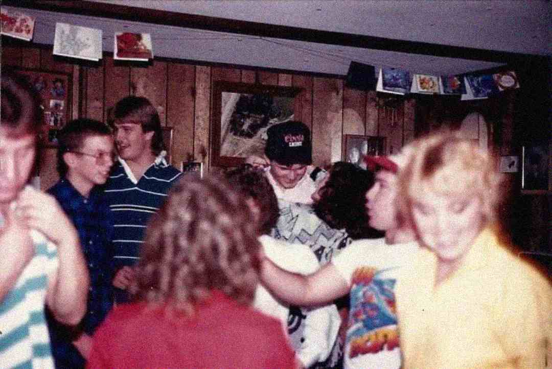 A group of teens gather indoors at a casual party, smiling and talking. The wood-paneled walls are decorated with cards and photos, and the scene is slightly blurry, giving a nostalgic, vintage feel.