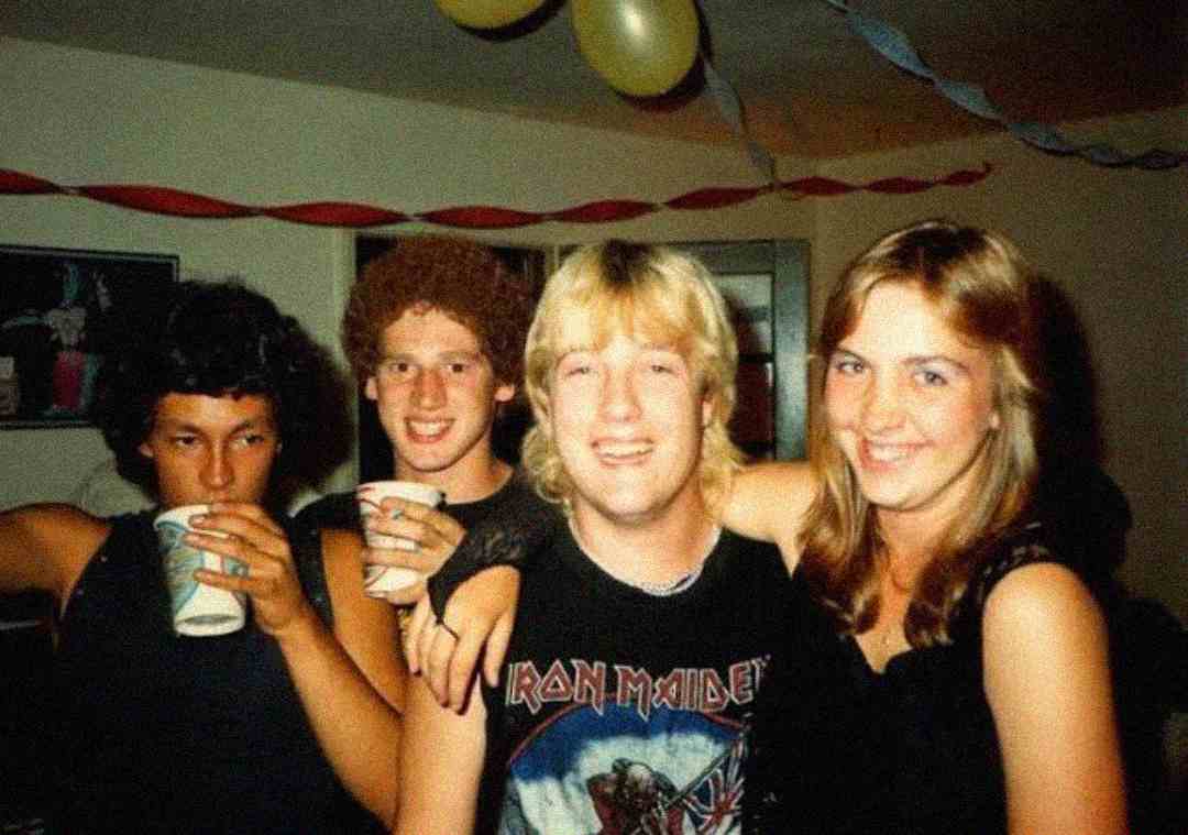 Four young adults at a party pose together, smiling. The person in front wears an Iron Maiden shirt. Two hold paper cups. Streamers and balloons decorate the room in the background.