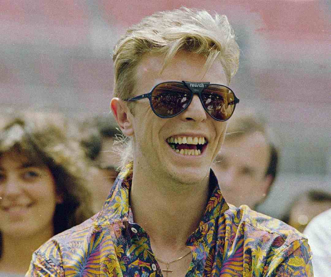 A person with blond hair smiles widely, wearing large dark sunglasses and a colorful patterned shirt, with people and a blurred background behind them.
