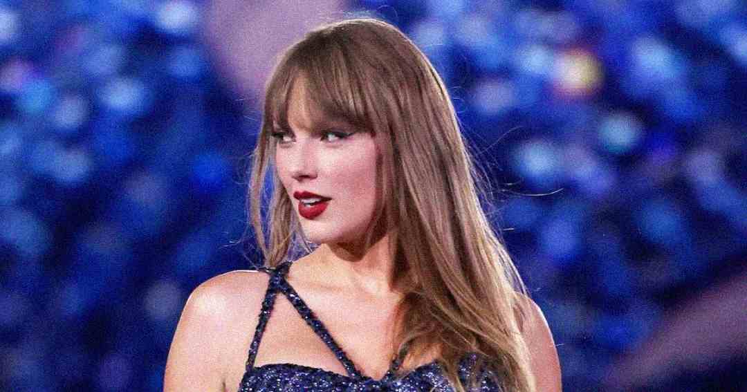 A woman with long, light brown hair and bangs is wearing a dark, sparkly top. She stands against a blurred blue background, looking slightly to the side with a confident expression.