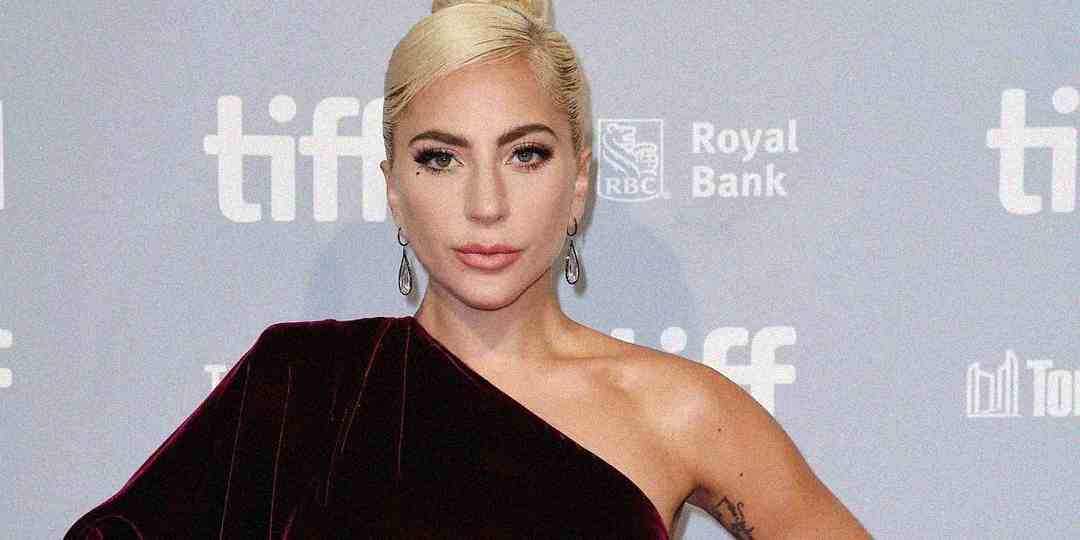 A person with platinum blonde hair in an elegant updo wears a dark, one-shoulder velvet dress and dangling earrings, posing confidently in front of a backdrop with "tiff" and sponsor logos.