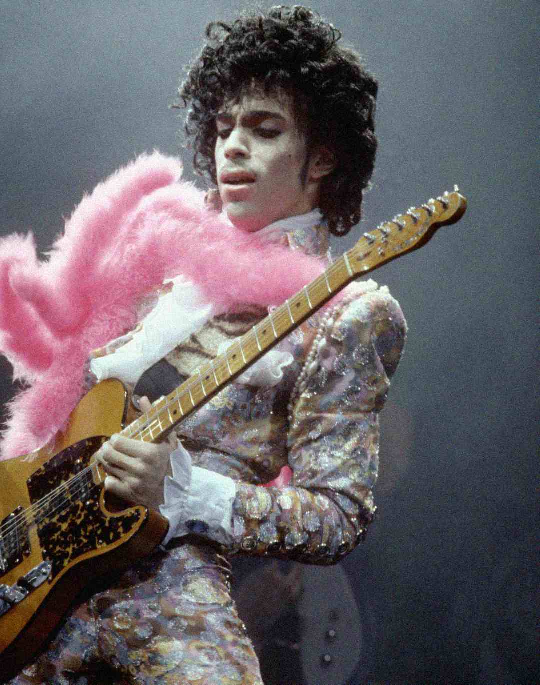 A musician with curly hair wears a patterned suit and a pink feather boa, playing an electric guitar on stage under dramatic lighting.