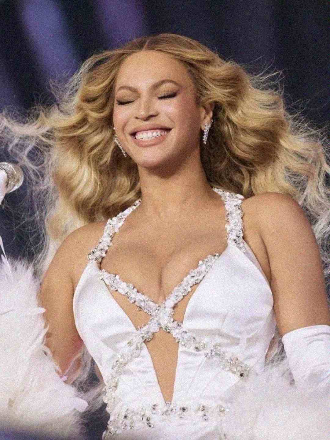 A woman with long, wavy blonde hair smiles with her eyes closed while performing on stage. She wears a glamorous white dress adorned with crystals and feathers, and stands in front of a microphone.