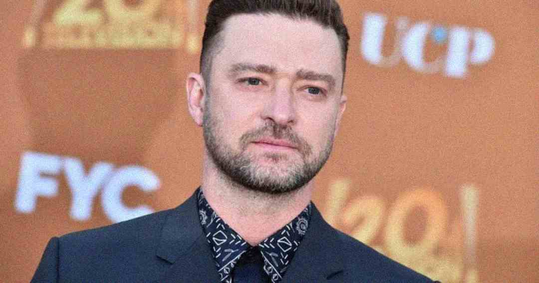 A man with short brown hair and a trimmed beard, wearing a dark suit and patterned shirt, stands in front of an orange background with blurred white and yellow text.