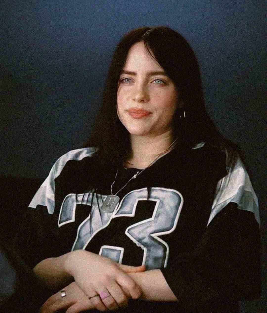 A young woman with long dark hair sits with her hands folded, wearing a black and white sports jersey with the number 23 and a few necklaces, against a dark background.