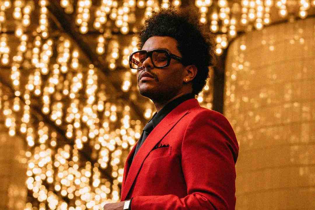 A man in a red suit and dark glasses stands under warm, glowing string lights, looking confidently to the side. The background is filled with blurred golden lights.