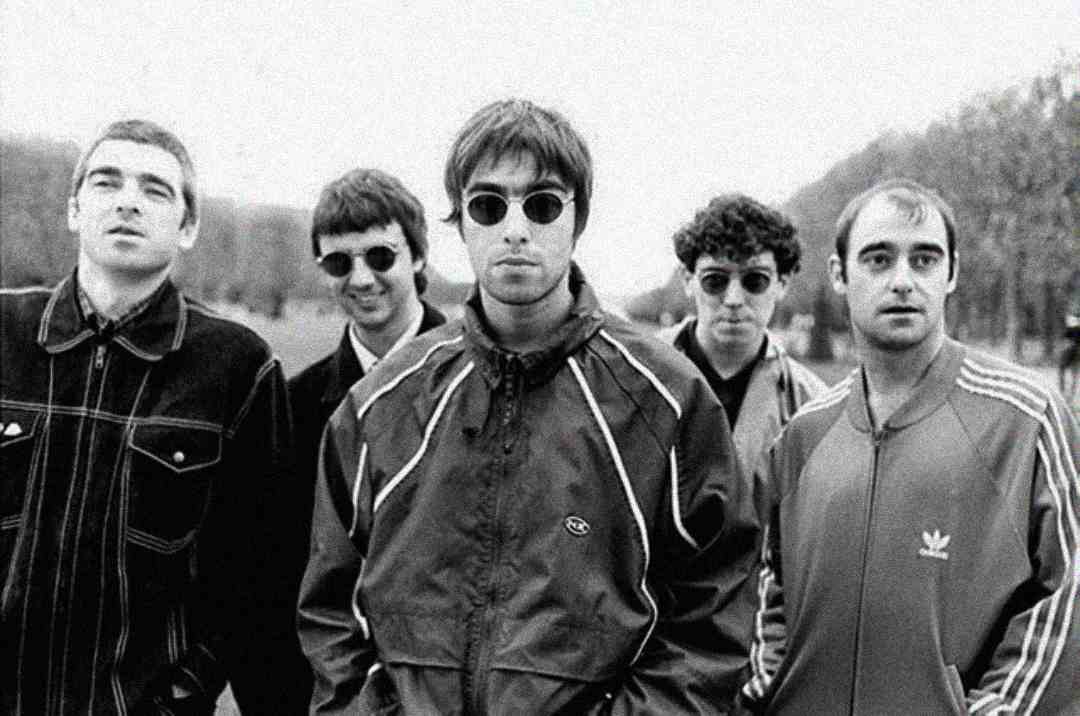 Five young men stand outdoors in a row, wearing casual jackets and sunglasses. The black and white photo has a vintage look, and trees line the background on either side of a path.
